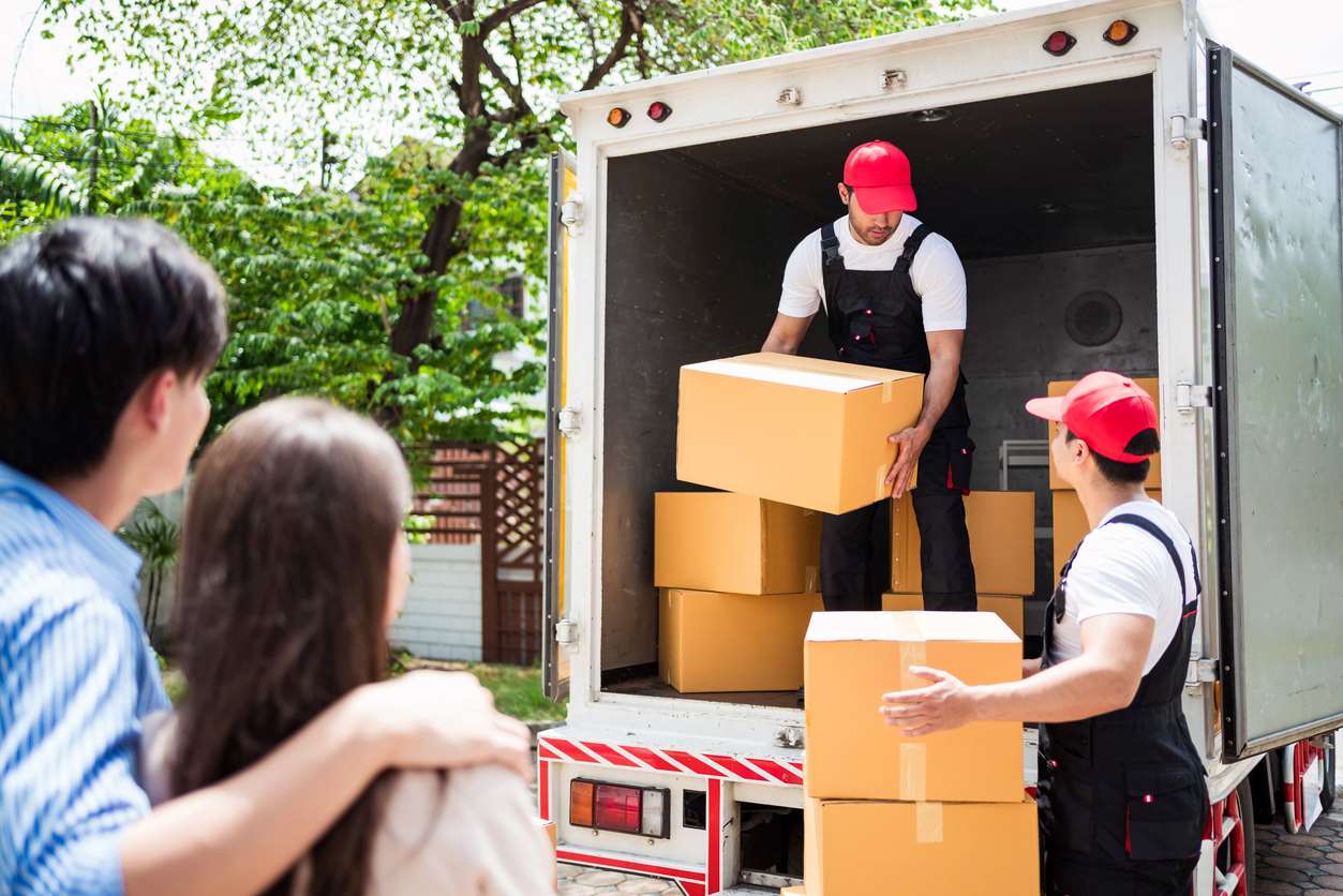 Man and woman watch as two men working for a moving company takes boxes out of a moving truck.