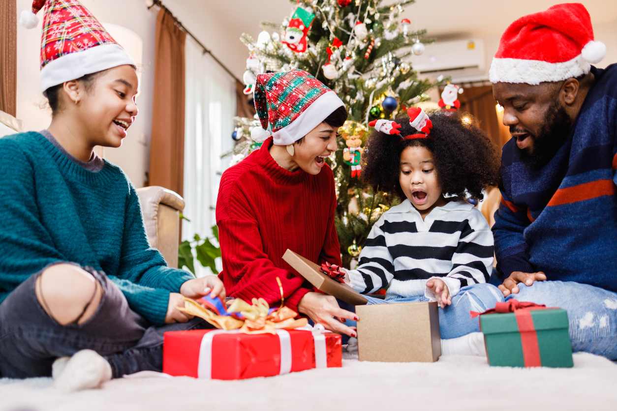A family in holiday clothing sits together as a young child opens a gift box