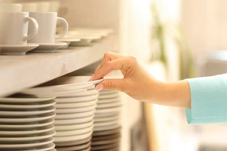 A woman in a blue sweater picks up a plate from a stack in the kitchen