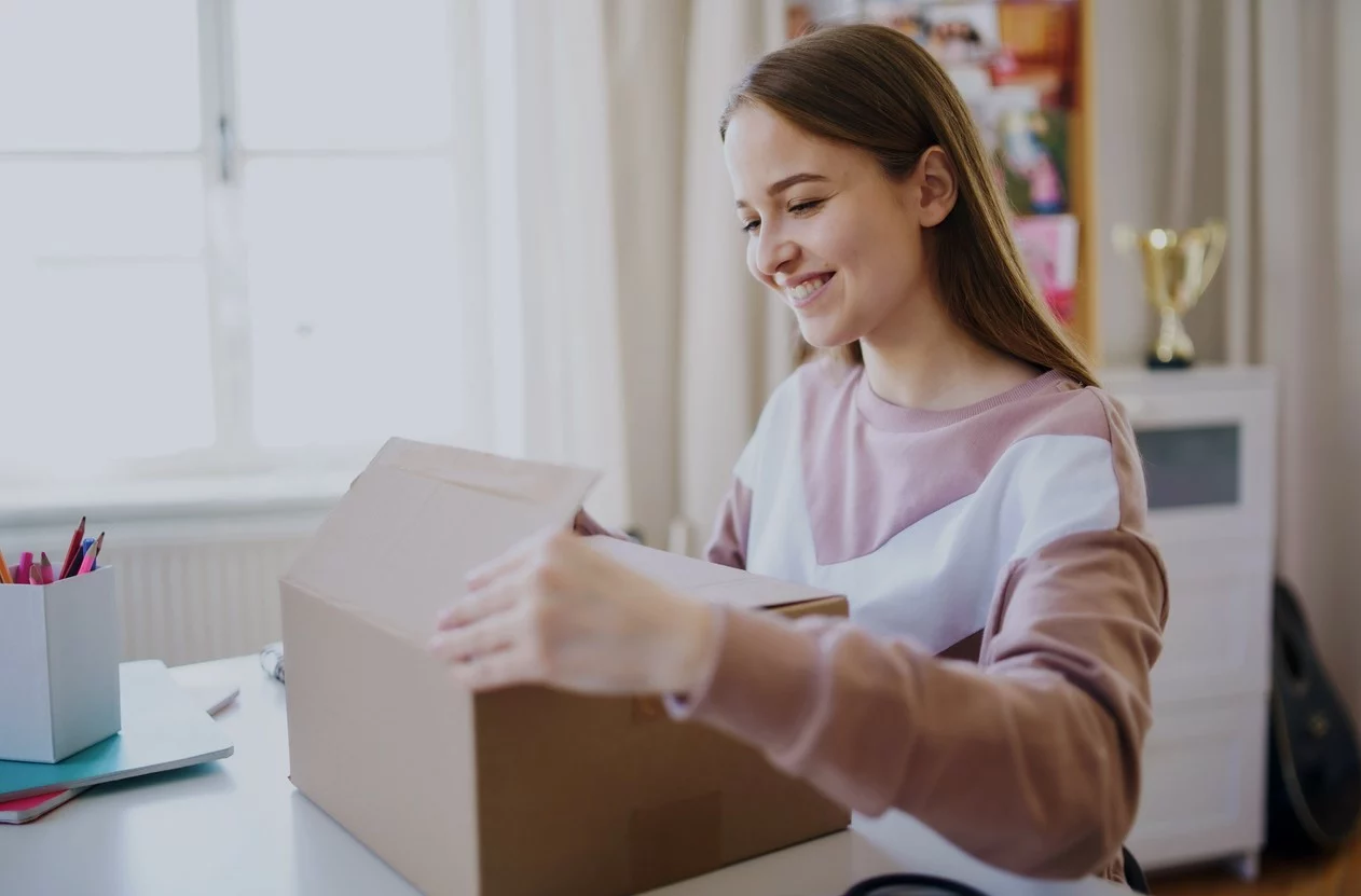 A student sits at a desk in a dorm room with a box of supplies in front of her