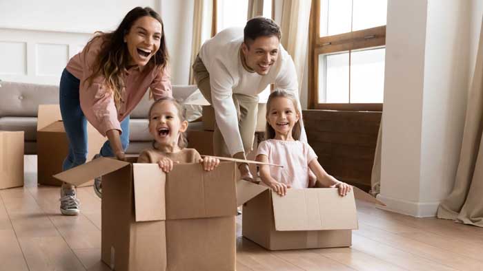 Smiling family. Parents are pushing two kids across the floor in cardboard moving boxes.