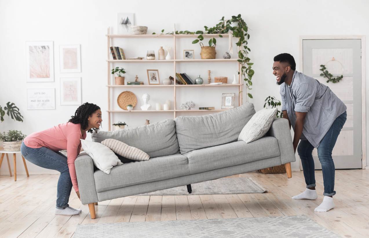 A man and woman move a modern gray colored couch into a bright living room space.