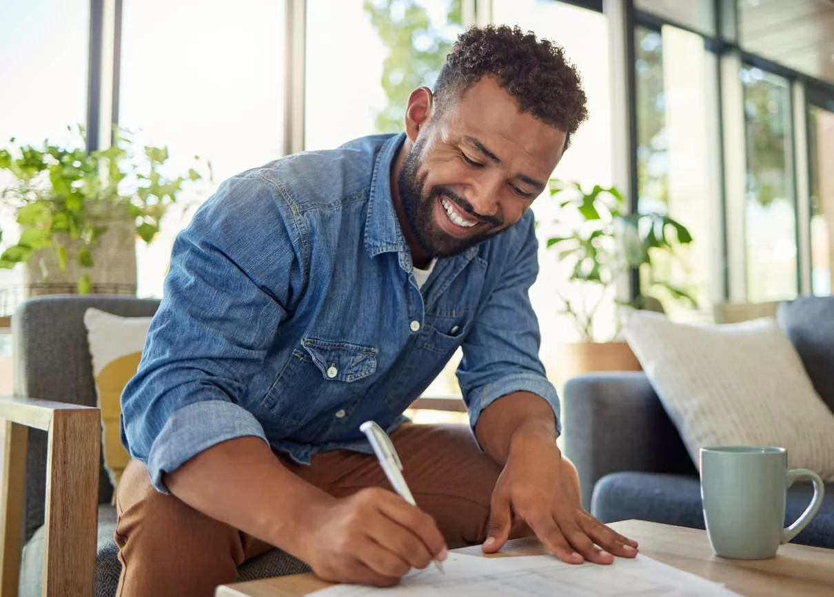 A man sitting in a gray chair uses a pen and smiles as he writes on a piece of paper