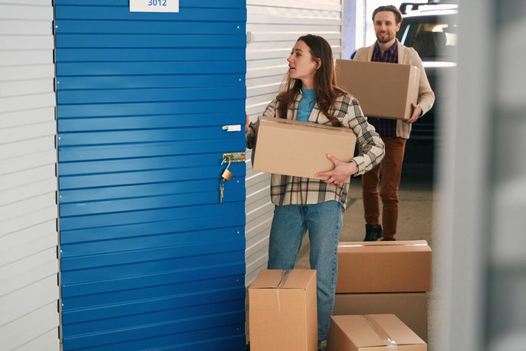 A man and woman carry cardboard storage boxes to a storage unit with a blue door. 