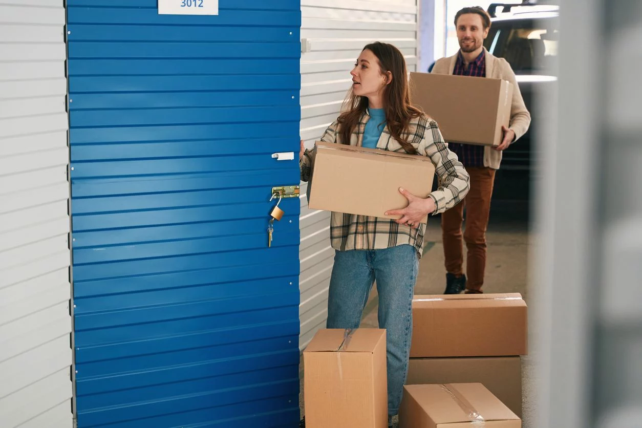 A man and woman carry cardboard storage boxes to a storage unit with a blue door. 
