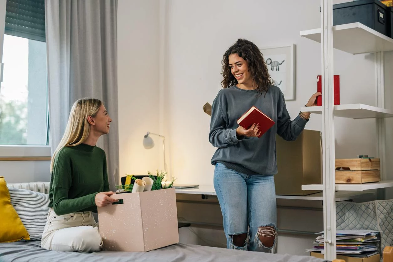 Two roommates sit in a dorm room while they decorate their space