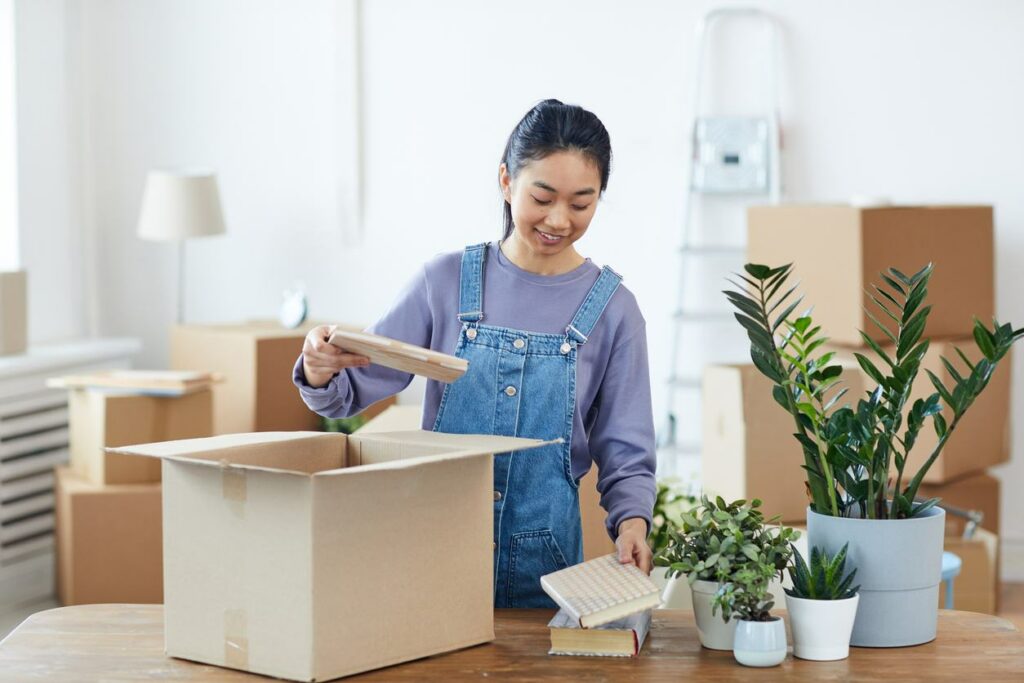 A woman stands at a table and packs books into a cardboard box. Plants sit next to her on the table.