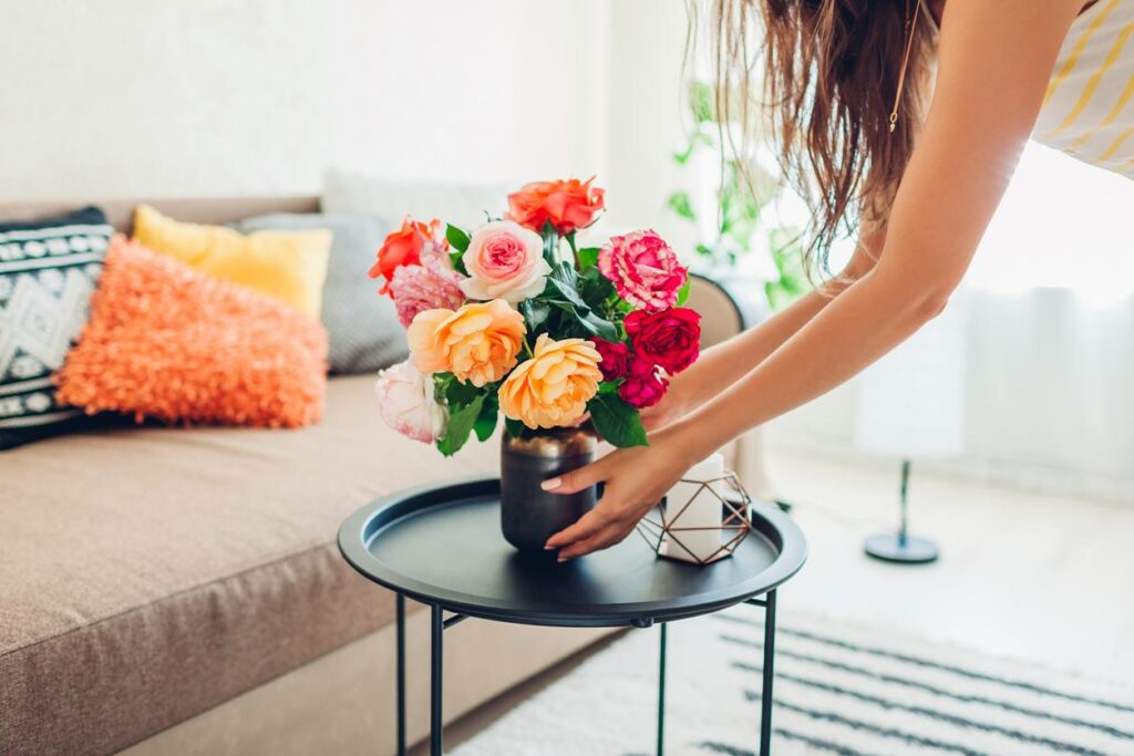 A woman sets a vase with pink and orange flowers on a black table in front of a brown colored couch. The room is brightly lit with natural light