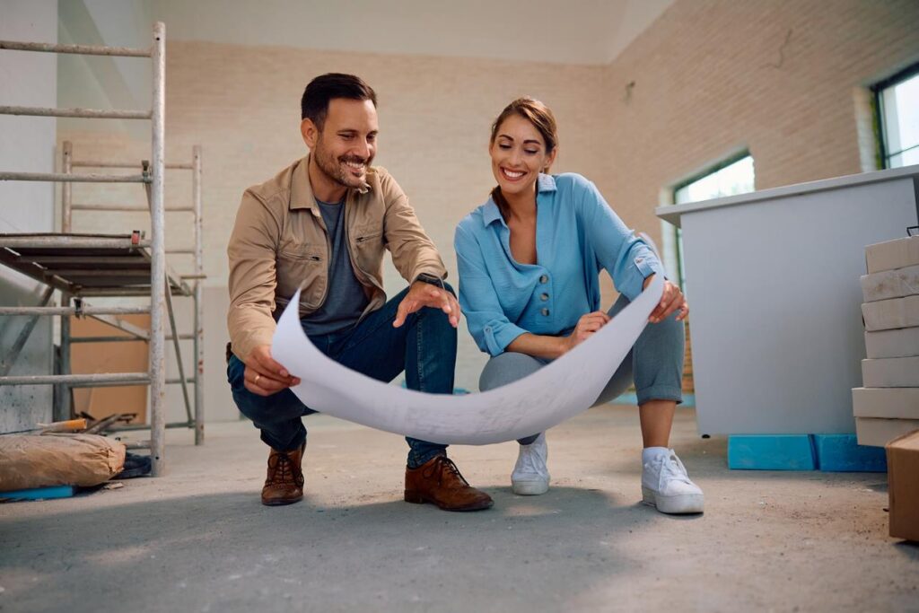 A happy couple holding blueprints inside a bare room for their spring home renovations.