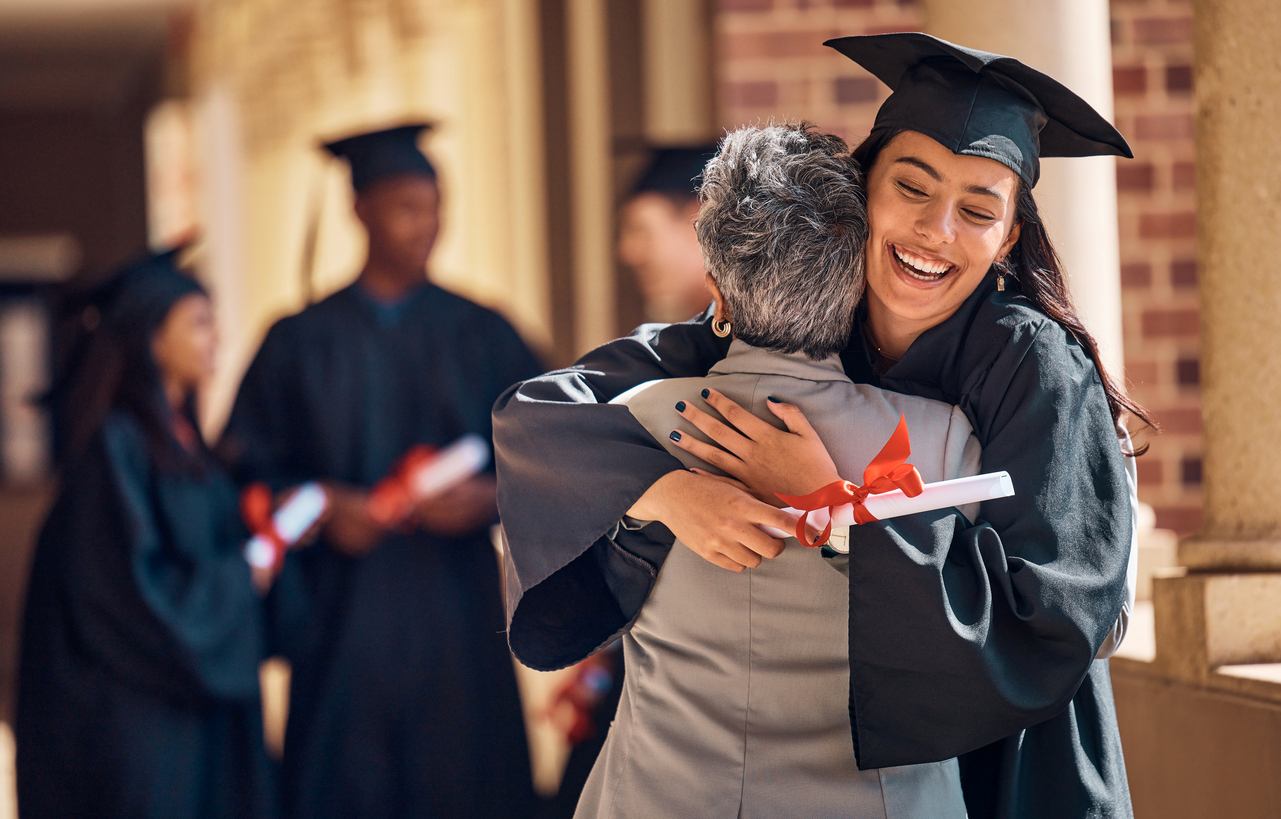 A happy college graduate hugs a family member in her cap and gown, holding a paper wrapped with red ribbon.