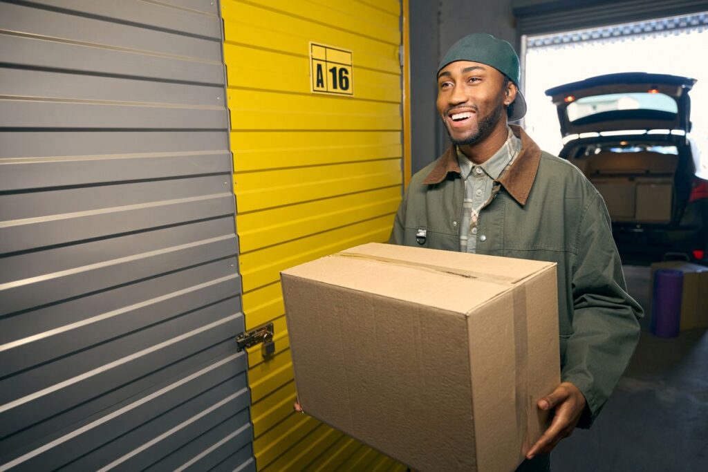 A smiling young man holding a cardboard box walks through a storage facility with an SUV with an open trunk behind.