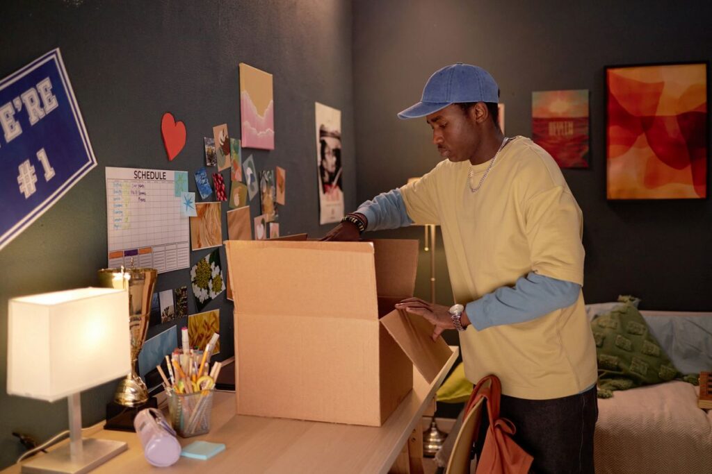 A college graduate in a yellow shirt and blue hat packs up his dorm by placing items in a cardboard box on a desk.
