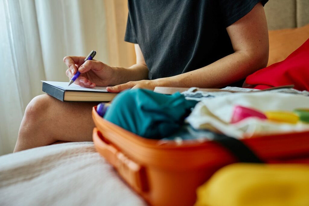 A person sitting on a bed next to an overflowing orange suitcase writes on a piece of paper with a pen.